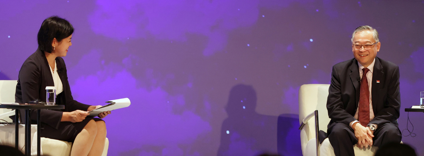 Two people seated on a stage; woman holds papers, man in a suit smiles, against a purple backdrop.